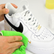 A person uses a green microfiber cloth and the Kleva Range “Make old shoes look new! Kleva Clean Sneaks Automatic Shoe Cleaner” to freshen up white Nike sneakers with a black swoosh and a small gold swoosh near the toe.