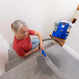An older woman with gray hair in a ponytail, wearing a red shirt, uses the Kleva Range KRAPOF® Carpet Shampoo Spray and hose attachment to clean a light gray carpet on a staircase.