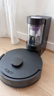 The robot on its dock on a timber floor, beside a sheer curtain.