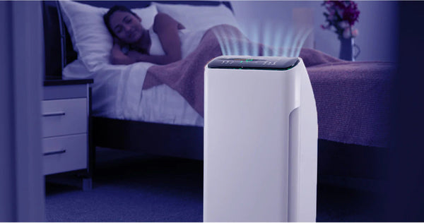 A white Kleva Range Remedical® Active Air Purifier with 7-stage filtration purifies the bedroom air at night, its blue lights aglow as a person sleeps under a brown blanket, ensuring effective odour and dust removal for restful sleep.