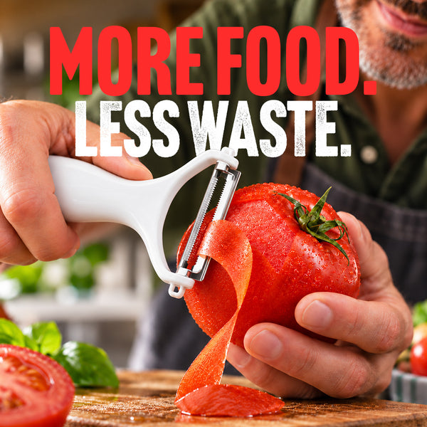 A person uses the Kleva Range Precision Peeler With Ultra Sharp Double-Sided Stainless Steel Blades to peel a tomato. The text above reads “MORE FOOD. LESS WASTE.” Tomatoes and herbs are scattered on a wooden surface in the background.