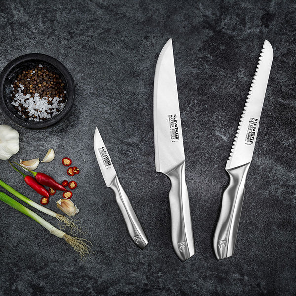 Three stainless steel knives from the Kleva Range KlevaCut® Master Series 3pc Professional Chef Knife Set are displayed on a dark gray countertop beside salt, pepper, fresh garlic, green onions, and sliced red chili peppers.