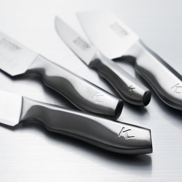 A close-up of the Kleva Range KlevaCut® LIMITED EDITION 4 Piece Premium Knife Set, featuring Japanese Vamolcrium steel blades and modern handles with a stylized K logo, displayed on a metallic surface. Includes BONUS bamboo case.