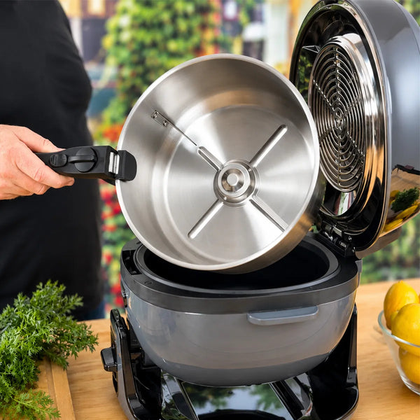 Someone lifts a stainless steel pot with a black detachable handle from an open air fryer or multi-cooker on the kitchen counter. Fresh herbs, lemons, and the KLEVA® Spin Crisp Cooking Accessories Set by Kleva Range are nearby.
