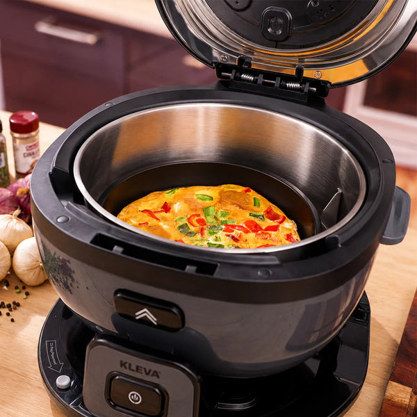 A Kleva Range multi-cooker sits open on a kitchen counter with a vegetable omelette in its metal pot, surrounded by garlic, a red spice jar, and the KLEVA® Spin Crisp Cooking Accessories Set.