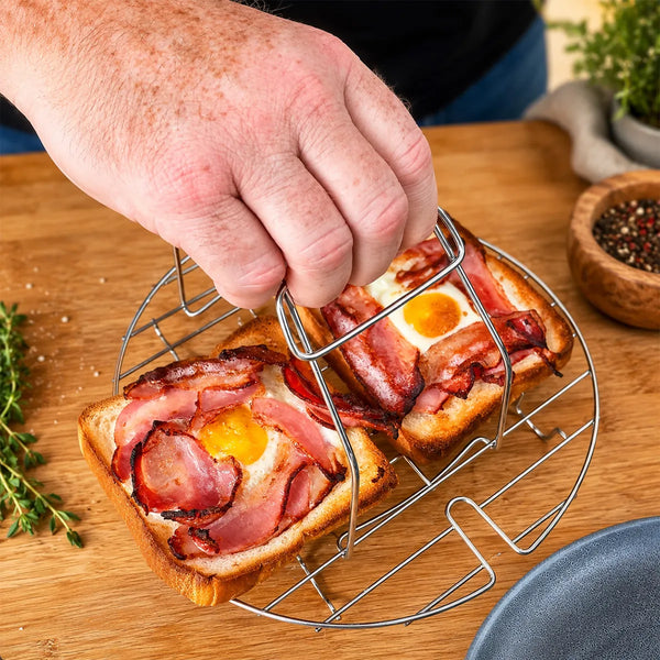A hand lifts the KLEVA® Spin Crisp Cooking Accessories Set by Kleva Range, its detachable handle holding a metal grill tray with two toast slices topped with bacon and baked eggs on a wooden surface, herbs and pepper bowls in the background.