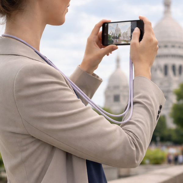 A person in a beige blazer uses their smartphone, secured with the KLEVA® Charge & Go Lanyard by Kleva Range, to photograph a domed building outdoors; the wearable phone charging cable hangs around their neck amid greenery on an overcast day.
