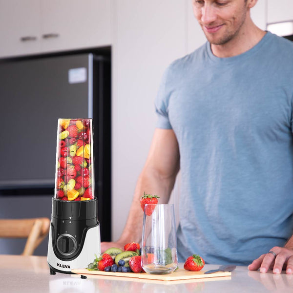 A man in a blue t-shirt stands in the kitchen beside a Kleva Range Health Hurricane GO™ Personal Blender with 700ml bottle, filled with fruit. A cutting board with berries and a glass are ready for quick, on-the-go smoothies anywhere.