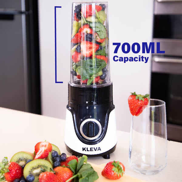 The Health Hurricane GO™ Personal Blender by Kleva Range, with a 700ml bottle, sits on a kitchen counter packed with spinach, strawberries, blueberries, and kiwi. Fresh fruit and spinach are nearby beside an empty glass garnished with a strawberry.