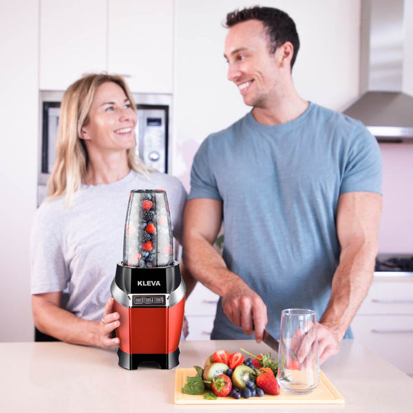 A woman stands by a red Kleva Range Health Hurricane Max™ Powerful 1000W Blender with Dual Cups, filled with fruit, smiling at a man slicing fruit in a modern kitchen. A glass and fresh fruits sit on the counter.