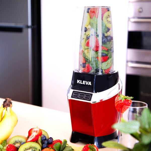 The Kleva Range Health Hurricane Max™ 1000W blender with dual cups sits on the counter, filled with fruits and spinach. Strawberries, blueberries, and bananas are scattered nearby, with a fridge and oven in the background.
