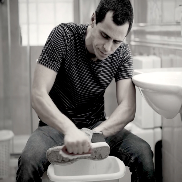 A man in striped t-shirt and jeans uses the Kleva Range "Kleva Clean Sneaks Automatic Shoe Cleaner" to refresh his sneaker over a plastic basin in a bathroom, surrounded by a sink, tiled walls, and bathroom essentials.