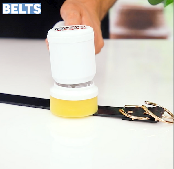 A person uses the Kleva Clean Sneaks Automatic Shoe Cleaner by Kleva Range from a white container to apply cleaner onto a black leather belt with a gold buckle on a white surface. "BELTS" is displayed in the top left corner.