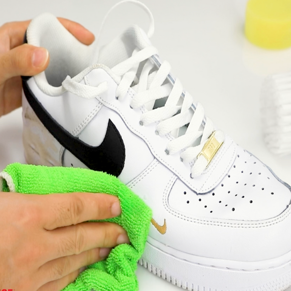 A person uses a green microfiber cloth and the Kleva Range “Make old shoes look new! Kleva Clean Sneaks Automatic Shoe Cleaner” to freshen up white Nike sneakers with a black swoosh and a small gold swoosh near the toe.