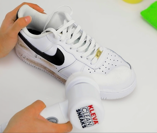 Someone is scrubbing a white sneaker with a black swoosh using the Kleva Range Make old shoes look new! Kleva Clean Sneaks Automatic Shoe Cleaner, while a green cloth rests in the background.