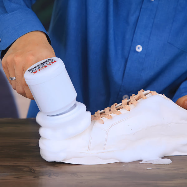 Wearing a blue shirt, someone uses the Kleva Range Kleva Clean Sneaks Automatic Shoe Cleaner to scrub a white sneaker, creating white foam on a wooden surface and making old shoes look new.