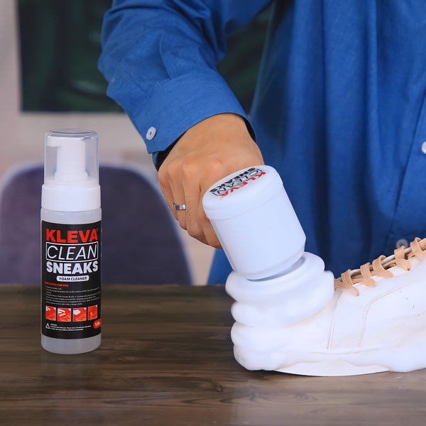A person in a blue shirt uses the Kleva Range Make old shoes look new! Kleva Clean Sneaks Automatic Shoe Cleaner to clean a white sneaker, with a bottle of Kleva Clean Sneaks on a wooden table beside them.