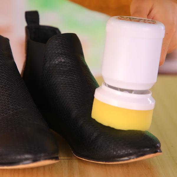 A hand uses the Kleva Clean Sneaks Automatic Shoe Cleaner by Kleva Range to clean and shine a black leather shoe on a wooden surface, while another black shoe sits beside the rechargeable device.