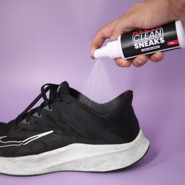 A hand sprays Kleva Range’s Make old shoes look new! Kleva Clean Sneaks Automatic Shoe Cleaner onto a black athletic sneaker with white soles, set against a plain light purple background.