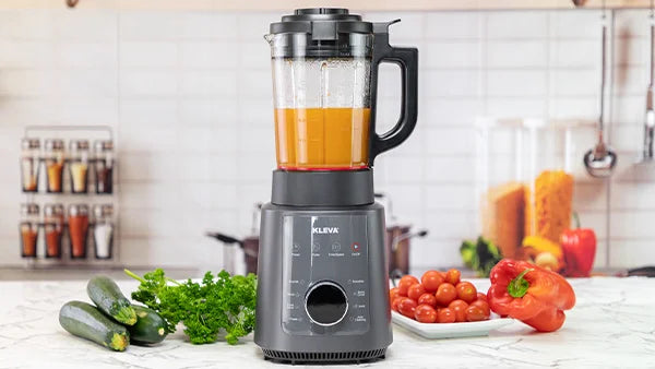 Kleva Mealio Blender with a sleek grey base and digital display, blending a vibrant orange soup alongside fresh zucchini, tomatoes, and red peppers.