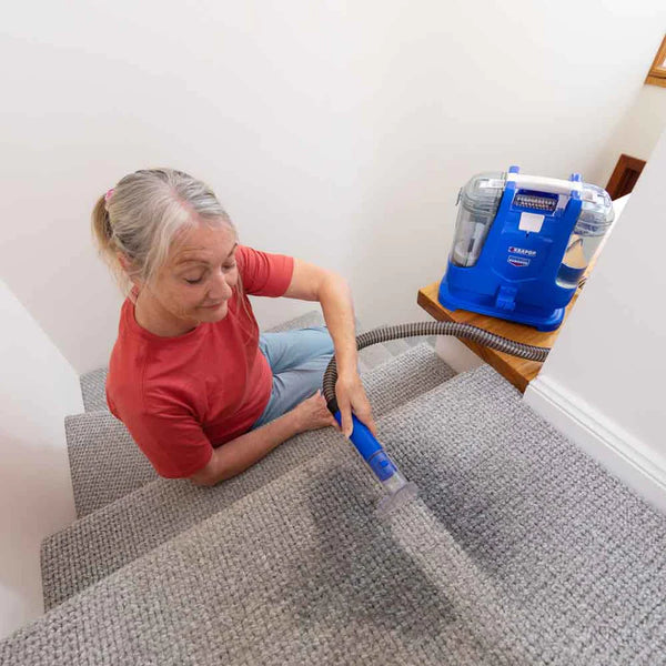 An older woman with gray hair in a ponytail, wearing a red shirt, uses the Kleva Range KRAPOF® Carpet Shampoo Spray and hose attachment to clean a light gray carpet on a staircase.