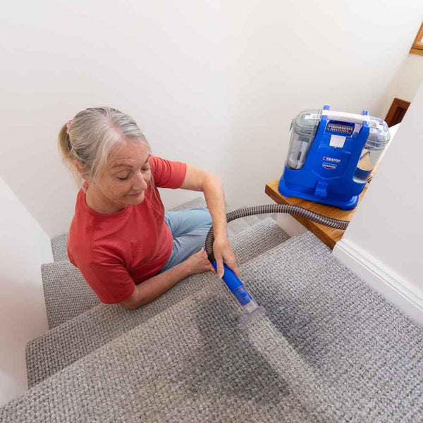 An older woman with gray hair tied back in a ponytail uses the Kleva Range KRAPOF® Spot & Stain Portable Remover with BONUS Cleaning Solution on a carpeted staircase, placing it on the step above as she tackles tough stains.