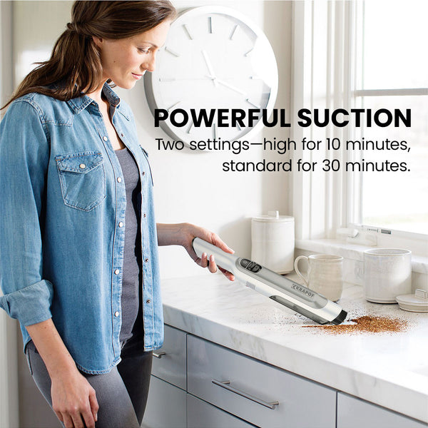 A woman uses the KRAPOF® Portable Mini Plus Handy Cordless Car Vacuum Cleaner by Kleva Range to clean spilled coffee grounds on a white kitchen counter. Text reads: POWERFUL SUCTION. Two settings—high for 10 min, standard for 30 min.