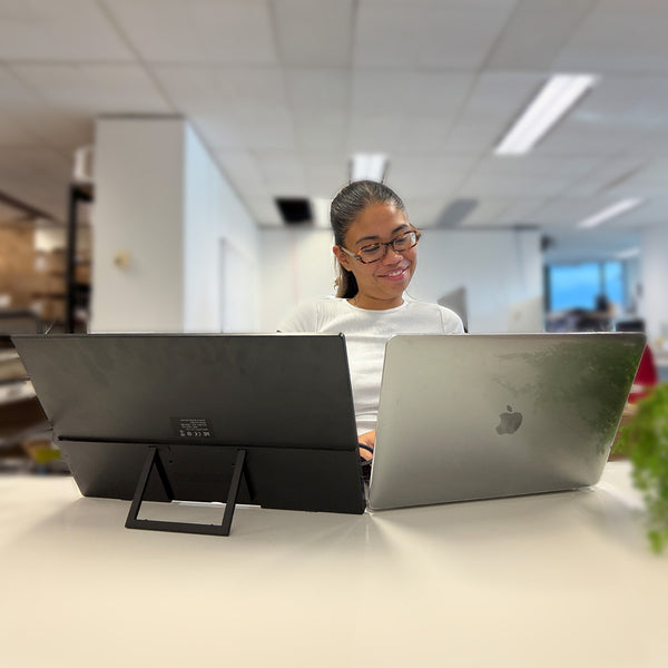 A person with glasses works at a desk using a laptop and the Kleva Range Kleva Portable 15.6" Screen Extender, a Full HD portable monitor, in a modern, well-lit office with blurred background details and a small plant on the right.