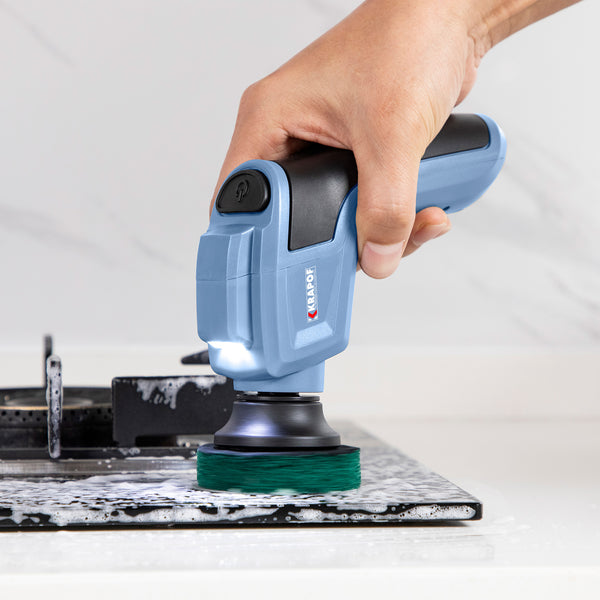 A hand uses the KRAPOF® Electric Lumi Scrub Pocket Cleaner by Kleva Range, featuring a blue handle, green pad, built-in LED light, and 5 replaceable heads, to easily clean a soapy, grimy stovetop.