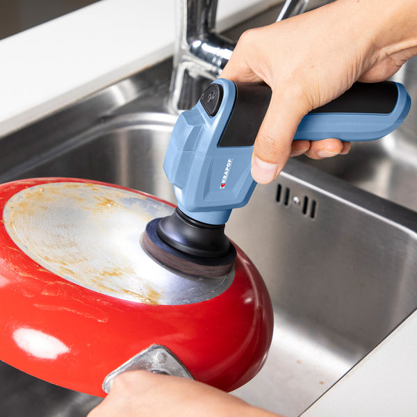 Using the KRAPOF® Electric Lumi Scrub Pocket Cleaner by Kleva Range with a round brush head, a person cleans the bottom of a red pan over a stainless steel kitchen sink.
