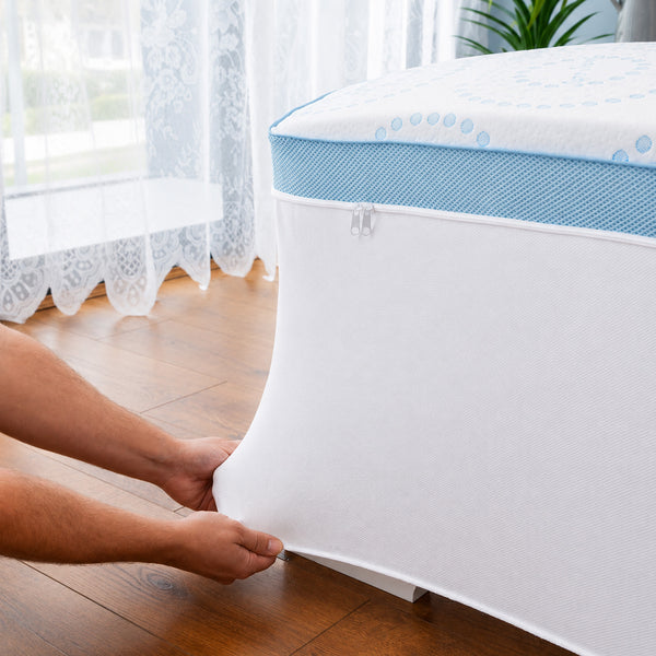 A person uses both hands to zip the Super Sleeper Pro Straight 2 Sleep® Mattress Topper with Cooling Technology onto a bed, which sits on a wooden floor near a window with sheer curtains and a potted plant. Two BONUS pillows are also included.