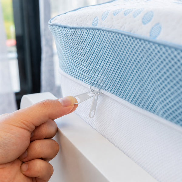 A hand pulls the zipper on a blue and white Super Sleeper Pro Straight 2 Sleep® Mattress Topper with Cooling Technology, placed next to a window with part of the mattress and bedding visible in the background, highlighting sleep comfort and innovative cooling features—plus 2 BONUS pillows.