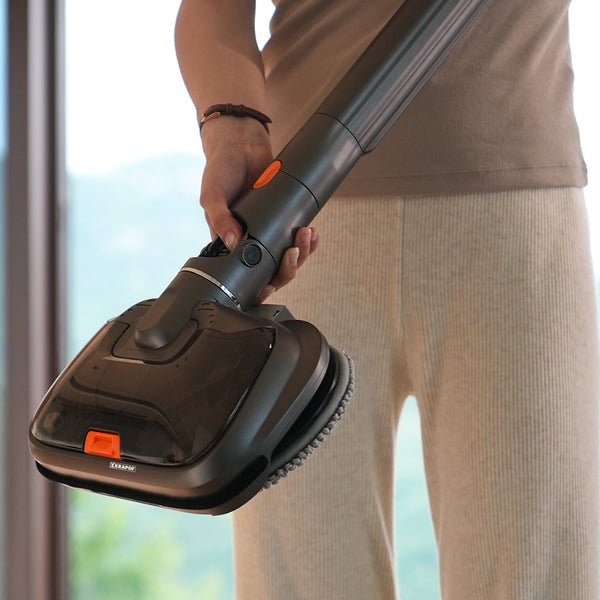 A person in beige pants and a brown top holds a Kleva Range KRAPOF® S3 Steam Drive Extension 2 in 1 Floor and Steam Mop for Q3 Stick Vacuum with a round microfiber pad near the camera; greenery is visible through a window in the background.