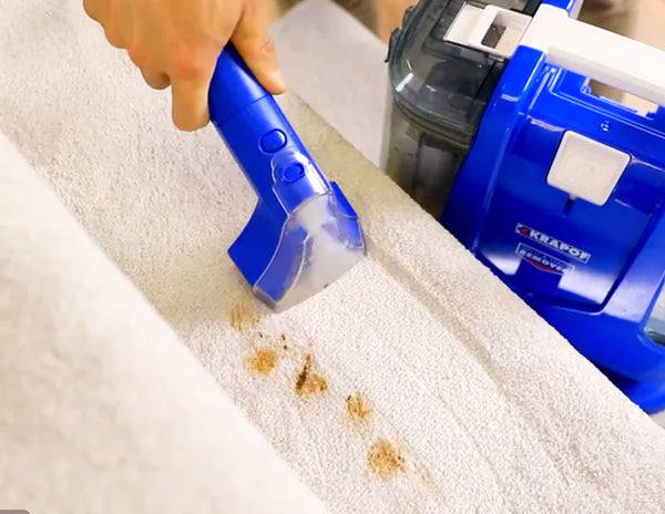 A person uses Kleva Range’s KRAPOF® Carpet Shampoo Spray to clean brown stains from a light-colored fabric. The stain remover is active, but some stains are still visible on the surface.