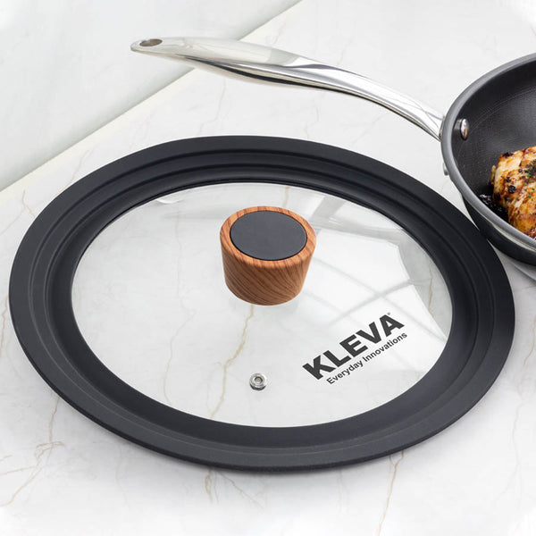 The Diamond Earth® Universal Glass Lid 24-28cm by Kleva Range, featuring a black rim and wooden handle, rests on a white countertop near a pan of cooked meat. This versatile lid fits pots between 24-28cm for all-purpose kitchen use.