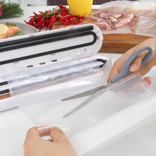 A person cuts a sheet from a Kleva® Vac Seal & Save Bag by Kleva Range near a vacuum sealer, with meat, chili peppers, and food storage bags visible on the kitchen counter.