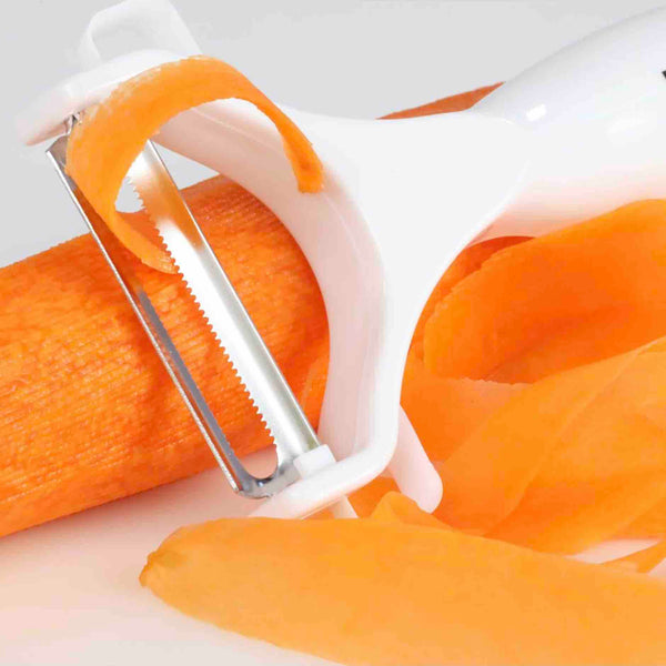 A Kleva Range z_archive_Kleva Safety Slicer™️ One-Push XL Vegetable Slicer + Sumo Slicer® peels a carrot, creating thin strips of skin around the blade and scattered on a white surface.