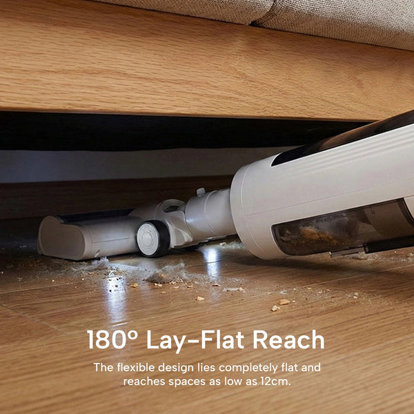 A cordless stick vacuum cleaner shown under a wooden bed or sofa, demonstrating its flexible design and reach. White and gray vacuum body with a transparent dust bin, placed on a wooden floor with debris, and text overlay reading '180° Lay-Flat Reach'.