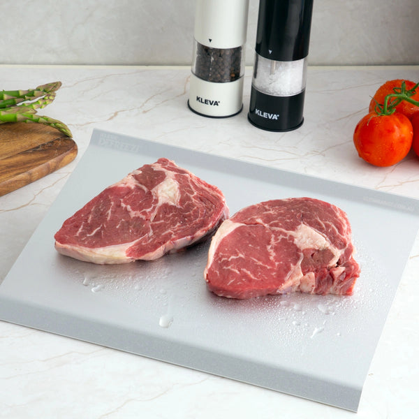 Two raw beef steaks rest on a cutting board atop a white countertop, ready to be expertly sliced using the KlevaCut® Master Series TV Special 4pc Knife Set with Knife Block & Defrosting Tray from Kleva Range. Nearby, salt and pepper grinders, a bunch of asparagus spears on a wooden board, and two ripe tomatoes await preparation. The gleaming Japanese stainless steel knives from the Kleva Range ensure every ingredient is cut with precision, making meal prep both effortless and stylish.