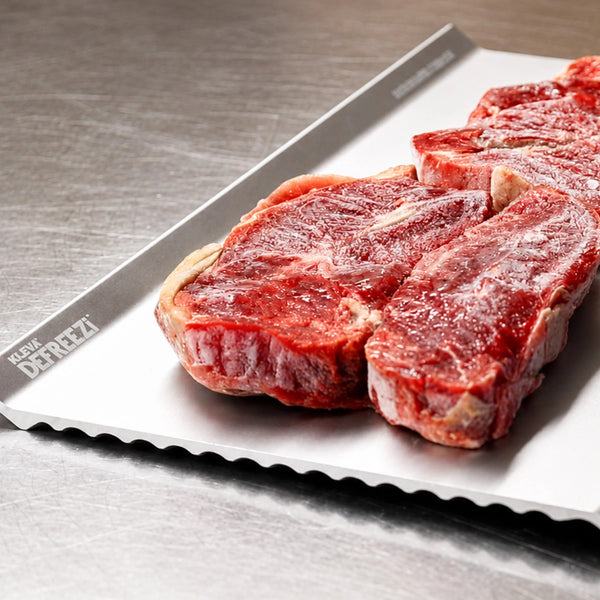 Three raw beef steaks rest on a metal defrosting tray labeled DEFREEZI in the corner, placed atop a stainless steel surface—perfectly complemented by the Kleva Range KlevaCut® Master Series TV Special 4pc Knife Set with Knife Block & Defrosting Tray, which features premium Japanese stainless steel knives for effortless slicing and preparation.