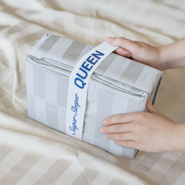 A pair of hands holds a folded, striped, light gray Imperial Dream Sheets™ 4pc Bedding Set by Super Sleeper Pro in luxurious 1200TC cotton sateen, wrapped with a white band labeled QUEEN, on a matching striped bed sheet.