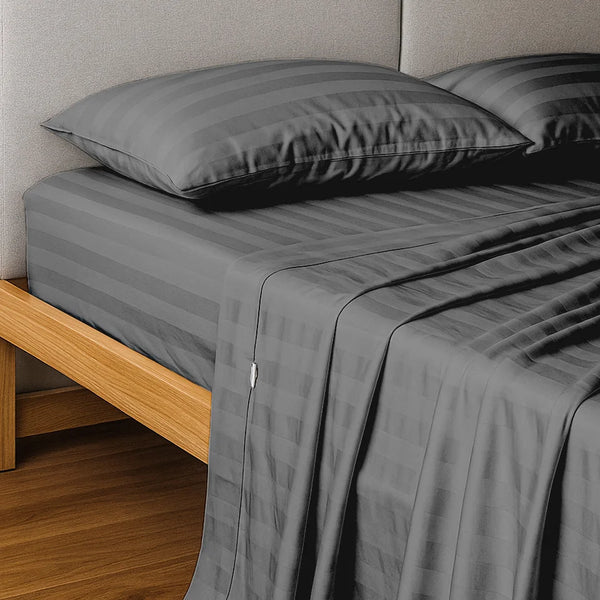 A neatly made bed features the Super Sleeper Pro Imperial Dream Sheets™ 4pc Bedding Set in dark gray stripes, 1200TC cotton sateen, with two matching pillows on a wooden frame against a light gray padded headboard.