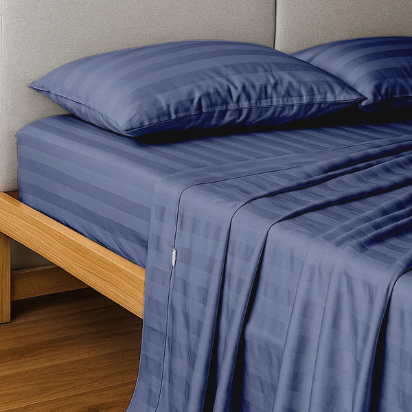 A neatly made bed features the Super Sleeper Pro Imperial Dream Sheets™ 4pc Bedding Set in blue stripes, crafted from 1200TC cotton sateen, with two pillows and a flat sheet on a wooden frame with gray upholstered headboard on a wooden floor.