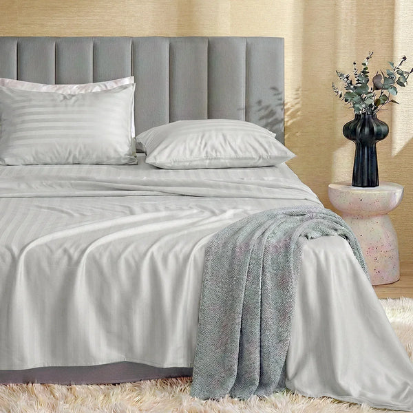 A neatly made bed features the Super Sleeper Pro Imperial Dream Sheets™ 4pc Bedding Set in light gray stripes, a gray upholstered headboard, textured gray blanket draped on the side, and a vase with greenery on a round side table.