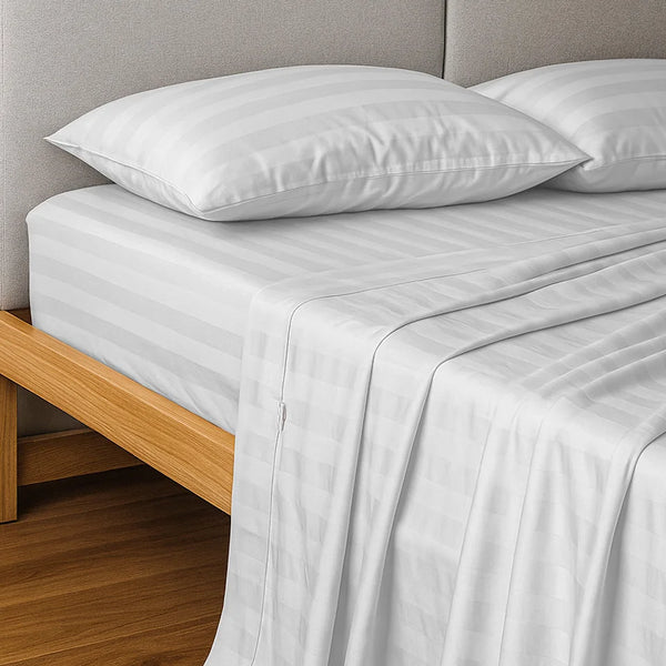 A neatly made bed showcases the Super Sleeper Pro Imperial Dream Sheets™ 4pc Bedding Set in light gray stripes, with two pillows and a wooden frame, set against a light gray upholstered headboard and wooden floor.