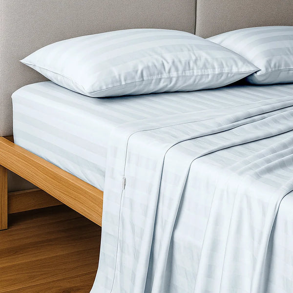 A bed features the Super Sleeper Pro Imperial Dream Sheets™ 4pc Bedding Set in white stripes, with two matching pillows on a light wood frame and beige upholstered headboard; secure-fit bedding drapes onto the wooden floor.