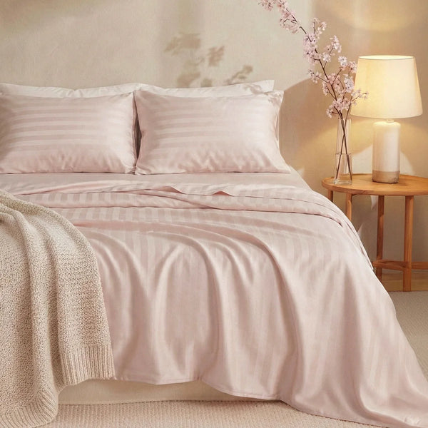 A neatly made bed features the Super Sleeper Pro Imperial Dream Sheets™ 4pc Bedding Set in light pink stripes, with two pillows, a cream knit blanket draped on one corner, next to a wooden nightstand with a beige lamp and pink flowers.
