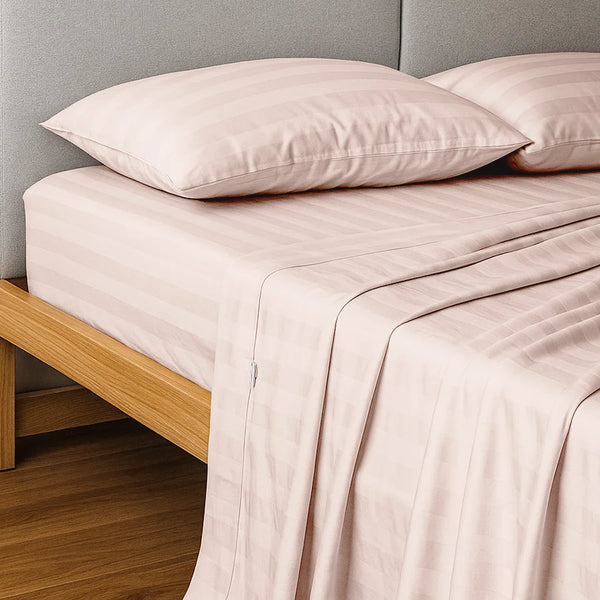 A neatly made bed features the Super Sleeper Pro Imperial Dream Sheets™ 4pc Bedding Set—1200TC cotton sateen with light pink stripes, fitted and flat sheets, two pillows—all atop a wooden frame and gray upholstered headboard.