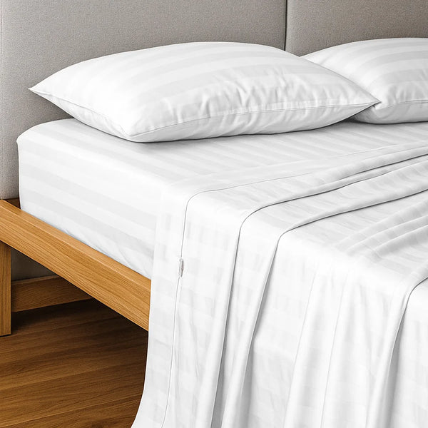 A neatly made bed features the Super Sleeper Pro Imperial Dream Sheets™ 4pc Bedding Set in white 1200TC cotton sateen, with two pillows on a wooden frame beside a light gray upholstered headboard and secure-fit bedding draped over the side.