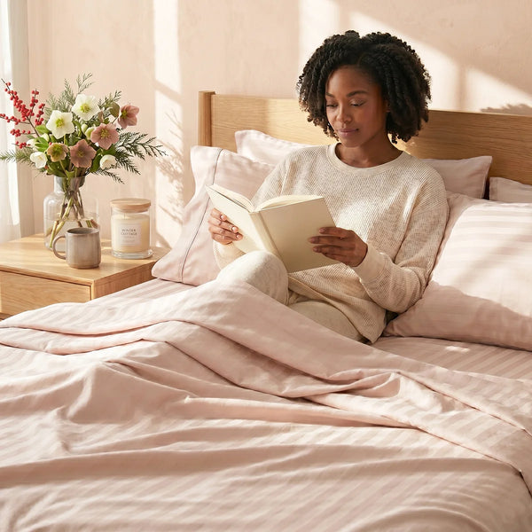 A woman sits up in bed reading, covered with Super Sleeper Pro’s Imperial Dream Sheets™ 4pc Bedding Set in light pink stripes, made from 1200TC cotton sateen. A wooden nightstand holds a vase with flowers and a candle in soft natural light.